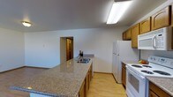 This single-floor space features a functional kitchen as its central highlight, designed with practicality and comfort in mind. The kitchen is outfitted with warm honey-toned wood cabinetry that provides ample storage, complemented by speckled granite-look countertops that extend generously, offering substantial workspace for meal preparation. A double-basin sink is integrated into the countertop, positioned conveniently along the counter run. White appliances, including a full-size refrigerator and a range with stovetop, lend a clean, cohesive look to the space. The kitchen layout follows an efficient configuration, with cabinetry lining the walls and an extended counter forming a natural divider between the kitchen and the adjoining living area. Light wood-tone flooring runs throughout, adding warmth and visual continuity across the space. Overhead, a large fluorescent light fixture provides bright, even illumination throughout the kitchen. The angled ceiling adds a subtle architectural detail, giving the space a slightly dynamic feel. A doorway visible in the background connects to additional areas of the home, hinting at a well-connected, open floor plan throughout.
