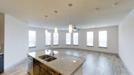 Located in Frisco, TX, this single-story space showcases a thoughtfully designed open-concept layout that seamlessly connects the kitchen and living areas. The kitchen is a standout feature, centered around a large island topped with granite countertops and equipped with a double basin sink and a modern pull-down faucet. Dark cabinetry beneath the island provides a striking contrast to the light stone surfaces, while pendant lighting above adds focused illumination and a contemporary aesthetic touch.

The adjoining living area is bright and welcoming, benefiting from a generous arrangement of tall windows that line the walls and invite abundant natural light throughout the day. A ceiling fan is centrally positioned to ensure comfortable airflow across the open space. The flooring transitions between large-format tile in the kitchen and wood-look flooring in the living area, defining each zone while maintaining a cohesive, modern look.

The neutral color palette of soft whites and grays throughout creates a clean, versatile atmosphere. The combination of open sightlines, quality finishes, and plentiful windows makes this Frisco property a well-appointed, light-filled space suitable for both everyday living and entertaining.
