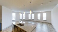 Located in Frisco, TX, this single-story space showcases a thoughtfully designed open-concept layout that seamlessly connects the kitchen and living areas. The kitchen is a standout feature, centered around a large island topped with granite countertops and equipped with a double basin sink and a modern pull-down faucet. Dark cabinetry beneath the island provides a striking contrast to the light stone surfaces, while pendant lighting above adds focused illumination and a contemporary aesthetic touch.

The adjoining living area is bright and welcoming, benefiting from a generous arrangement of tall windows that line the walls and invite abundant natural light throughout the day. A ceiling fan is centrally positioned to ensure comfortable airflow across the open space. The flooring transitions between large-format tile in the kitchen and wood-look flooring in the living area, defining each zone while maintaining a cohesive, modern look.

The neutral color palette of soft whites and grays throughout creates a clean, versatile atmosphere. The combination of open sightlines, quality finishes, and plentiful windows makes this Frisco property a well-appointed, light-filled space suitable for both everyday living and entertaining.