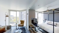 Located at 1101 East Bayaud Avenue in Denver, CO, this contemporary high-rise unit sits within the desirable Washington Park neighborhood, offering an elevated urban living experience. The open-concept layout is anchored by expansive floor-to-ceiling windows on multiple sides, flooding the interior with abundant natural light while framing panoramic views of the Denver skyline, neighboring rooftops, and lush tree canopies below.

The living area is thoughtfully appointed with a modern aesthetic, featuring clean white ceilings, light hardwood-style flooring, and a curated mix of contemporary furnishings. A plush seating arrangement with a leather accent chair and cowhide rug creates an inviting lounge space, perfectly positioned to take advantage of the sweeping corner views. A flat-screen television mounted on a sleek media console bridges the living and sleeping areas seamlessly.

The sleeping area is defined by an elegant four-poster bed frame with crisp white bedding, complemented by soft ambient lighting. The open, airy design maintains a sense of continuity throughout the space, while the corner window configuration ensures natural light reaches every corner. The overall ambiance is sophisticated yet comfortable, blending modern design sensibilities with the warmth of a well-curated interior — ideal for those seeking a refined urban retreat in the heart of Denver.