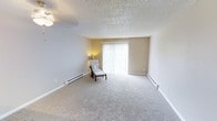 This single-floor, 1-bedroom, 1-bathroom residence offers a well-organized and efficient layout designed for comfortable everyday living. The living area is finished with plush wall-to-wall carpeting in a neutral tone, contributing to a warm and inviting atmosphere throughout the space. A textured ceiling adds subtle architectural character, while clean white baseboards frame the room with a crisp, finished look. A sliding glass door fitted with vertical blinds serves as the focal point of the living area, allowing abundant natural light to fill the room and providing access to the outdoors. Baseboard heating runs along the perimeter of the living space, ensuring consistent warmth during cooler months. The neutral wall palette in soft beige and white tones creates a versatile backdrop well-suited for a variety of interior styles. The overall ambiance of this residence is calm and understated, offering a practical and cozy environment. Its compact yet thoughtfully arranged footprint makes efficient use of every square foot, making it an accessible and easy-to-maintain living space.