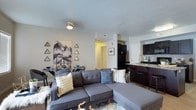 This single-floor residence features an open-concept layout that seamlessly integrates the living, dining, and kitchen areas into one cohesive space. The living area serves as the central hub, anchored by a spacious gray sectional sofa accented with decorative pillows. A textured area rug and geometric wall art featuring triangular mountain motifs add visual interest and a modern touch to the neutral-toned walls.

The dining area is positioned adjacent to the living space, accommodating a sleek table with seating for multiple guests, making it well-suited for both everyday meals and entertaining. Flowing naturally from the dining area, the kitchen is equipped with dark cabinetry, black appliances, granite-style countertops, and a breakfast bar with bar stools. A single-basin sink sits beneath upper cabinetry, maximizing storage and functionality.

The open layout promotes a sense of spaciousness throughout, enhanced by consistent flooring that runs across the main living areas. A hallway visible in the background leads to additional private areas of the residence. The overall ambiance is modern and well-appointed, with a neutral color palette complemented by contemporary décor and finishes.