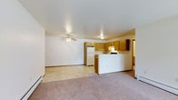 This single-floor space offers an open-concept layout that seamlessly connects the living area and kitchen, creating a functional and cohesive environment. The living area is finished with plush carpeting and features a ceiling fan with integrated lighting, providing both comfort and air circulation. A baseboard heating unit runs along the perimeter, ensuring efficient warmth throughout the space.

The kitchen is defined by warm honey-toned wood cabinetry that provides ample storage, complemented by a white refrigerator and a range. A white peninsula separates the kitchen from the living area, offering additional workspace and a natural division between the two zones while maintaining the open feel. The kitchen floor transitions to a light-toned vinyl or linoleum surface, practical for easy maintenance.

Overhead lighting in the kitchen adds to the bright, well-lit atmosphere of the space. A doorway visible beyond the kitchen suggests access to additional rooms, adding to the overall functionality of the layout. The neutral color palette of white walls and warm wood tones throughout creates a clean, inviting ambiance. The straightforward, practical design makes this space well-suited for everyday living.