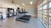 This modern fitness center offers a spacious, single-floor layout designed to support a full range of workout routines. The interior is finished with light wood-look flooring and neutral gray-toned walls accented by a contrasting horizontal stripe, creating a clean and contemporary aesthetic throughout.

The cardio zone is anchored by a row of Matrix treadmills and elliptical trainers, each equipped with individual display screens for an interactive workout experience. Ceiling fans provide additional air circulation alongside the recessed and flush-mounted fluorescent lighting overhead. A wall-mounted flat-screen television adds an entertainment element to the space.

The strength and functional training area includes a well-stocked dumbbell rack, a vertical knee raise station, stability balls, resistance mats, and a cable machine, offering versatile options for users of all fitness levels. A compact refrigerator near the entrance provides convenient access to refreshments.

Large garage-style windows and glass doors line the perimeter, flooding the interior with natural light and offering open views of the surrounding outdoor landscape, including palm trees and green spaces. The glass entrance door features posted fitness center guidelines, and the overall layout ensures ample floor space for free movement and stretching. The bright, airy environment makes this fitness center an inviting and functional destination for daily exercise.