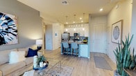 This single-floor residence offers a thoughtfully designed open-concept layout that seamlessly connects the living and kitchen areas, creating a bright and cohesive living environment. Light-toned wood-look flooring flows throughout, tying the spaces together with a clean, modern aesthetic.

The kitchen is a standout feature, boasting white cabinetry that extends to the ceiling with glass-front upper display cabinets, stainless steel appliances including a refrigerator with ice dispenser, microwave, and range, as well as a granite-topped peninsula with seating for two. Sleek pendant lighting hangs above the peninsula, adding both function and style. Neutral grey subway tile backsplash complements the overall color palette.

The living area is generously proportioned, allowing for comfortable furniture arrangements and accented by recessed lighting overhead. Warm, neutral wall tones create an inviting ambiance throughout. A hallway corridor leads to additional rooms, and the layout provides practical flow between all areas. The space is finished with ceiling-height doors and visible HVAC vents, reflecting attention to both comfort and detail. Overall, the residence presents a clean, contemporary atmosphere well-suited for modern living.