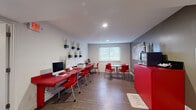 This single-floor business center is a compact yet well-organized communal workspace designed to accommodate productivity and casual collaboration. The room features a striking red-and-white color scheme that gives the space a modern, energetic character. Along one wall, a long red workstation desk hosts multiple iMac computers alongside a printer, providing ample workspace for residents or guests needing access to technology. Bold red chairs complement the desk throughout the space, maintaining a cohesive design aesthetic.

A round table with matching red chairs is positioned near a window fitted with horizontal blinds, allowing natural light to filter into the room and creating an inviting spot for meetings or informal gatherings. Colorful abstract artwork adorns the accent walls, adding visual interest against the contrasting light and dark gray wall tones.

A dedicated coffee station, complete with a coffee maker and cup supplies, is set atop a red cabinet, offering a convenient amenity for users of the space. The room is finished with warm wood-look flooring throughout, recessed ceiling lighting, and a wall-mounted whiteboard for note-taking. A hand sanitizer dispenser, decorative mirror, and wall sculptures complete the thoughtfully curated interior, making this business center both functional and visually engaging.