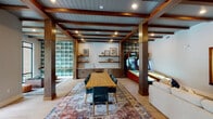 Located at 5824 McCrimmon Pkwy in Morrisville, NC, this thoughtfully designed single-floor amenity space blends modern style with inviting warmth. The interior is anchored by rich wood beam ceilings with shiplap detailing, bold teal and deep blue accent walls, and light hardwood flooring that flows throughout. A striking set of glass-paneled black double doors welcomes visitors into the main area, immediately revealing the space's open, airy layout. The game room is a standout highlight, featuring exposed wood beam ceilings, a skeeball arcade machine, a wall-mounted Scrabble board, a large communal dining table with upholstered chairs, and a plaid-tiled accent wall. A built-in wet bar with hexagonal tile backsplash and open shelving provides a convenient refreshment station. An adjacent lounge area features a sectional sofa, a textured 3D wood panel feature wall, built-in bookshelves, and a cozy seating nook. A bar-height counter with teal upholstered stools sits beside floor-to-ceiling windows overlooking an outdoor pool and patio area, filling the space with natural light and scenic views. Black-framed French doors offer direct access to the pool deck, seamlessly connecting indoor and outdoor living.