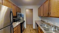 This single-floor space offers a practical and comfortable layout designed for everyday living. The kitchen is a standout feature, designed in a classic galley configuration that maximizes efficiency. Warm-toned wood cabinetry lines both sides of the kitchen, providing generous storage space with upper and lower cabinets throughout. The countertops feature a granite-look laminate surface, offering a functional workspace for meal preparation. Stainless steel appliances include an electric range with a built-in microwave above, and a dishwasher conveniently positioned near the double-basin stainless steel sink. Light wood-look flooring runs the length of the kitchen, adding warmth and easy maintenance to the space. A textured ceiling adds a traditional touch to the overall aesthetic. A doorway at the end of the kitchen connects seamlessly to the adjoining living area, creating a natural flow throughout the floor plan. The neutral wall tones throughout keep the space feeling bright and open, providing a versatile backdrop for a variety of interior styles. Overall, the space presents a clean, functional design well-suited for comfortable day-to-day living.