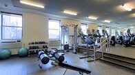 This fitness center is designed to support a comprehensive workout experience within a single, open-plan floor. The space is illuminated by a combination of overhead fluorescent lighting and natural light streaming through multiple windows, creating a bright and energizing environment. The walls are finished in a neutral tone, complemented by a large mirror along one side that enhances the sense of depth and allows users to monitor their form during exercise. The flooring consists of durable, speckled rubber or composite material suited for heavy equipment use. The strength training area features a multi-function cable machine and a dumbbell rack stocked with a range of free weights, alongside an adjustable workout bench for versatile strength exercises. The cardio section is equipped with multiple treadmills, providing ample options for aerobic training. A wall-mounted air conditioning unit ensures climate control throughout the space. The overall layout is organized and functional, with equipment arranged to allow ease of movement between stations, making the facility suitable for a variety of fitness routines.