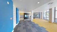 This dynamic single-floor commercial space combines modern design with functional versatility, making it well-suited for collaborative work or community activities. The open layout is visually striking, anchored by bold blue accent walls paired with a distinctive stone-textured feature wall that adds depth and character to the interior. Patterned carpet tiles in a mix of gray tones cover the main area, transitioning to a warm yellow flooring in an adjacent open zone, clearly delineating different functional areas within the space. A built-in kitchenette is seamlessly integrated into one wall, featuring warm wood cabinetry, a mosaic tile backsplash, a wall-mounted flat-screen television, and a beverage cooler — creating an inviting break or social area. Recessed lighting throughout provides even, modern illumination across the open floor plan. A structural column adds an architectural element to the space without interrupting the sense of openness. Large glass-paneled doors and windows at the far end of the space flood the interior with natural light while offering views into an adjacent area, enhancing the bright and airy ambiance. The overall atmosphere is energetic yet polished, reflecting a well-designed, contemporary environment.