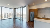 This contemporary high-rise residence offers a thoughtfully designed single-floor layout that balances modern aesthetics with functional living. The open-concept living area is anchored by dramatic floor-to-ceiling windows that frame sweeping panoramic city views, flooding the space with natural light and creating a strong visual connection to the urban skyline beyond.

The kitchen is a standout feature, outfitted with warm wood cabinetry at both upper and lower levels, complemented by a marble-style tile backsplash. A full suite of stainless steel appliances includes a gas range, built-in microwave, dishwasher, and a large French door refrigerator. A central island with a clean white countertop provides additional workspace and casual dining space.

Hardwood-style flooring runs throughout the residence, lending warmth and continuity across each area. The bedroom offers generous proportions, highlighted by floor-to-ceiling windows with city views, a ceiling fan, and a sliding barn door that opens to a well-organized walk-in closet.

The bathroom is finished with a wood-framed vanity mirror, white quartz countertop, wood-toned cabinetry, and a tiled walk-in shower. Clean white walls and recessed lighting throughout maintain a bright, minimal atmosphere that complements the residence's modern character.