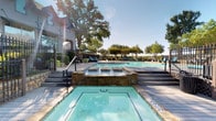 This outdoor pool area offers a well-appointed, resort-style retreat designed for relaxation and recreation. The centerpiece is a generously sized swimming pool with crystal-clear turquoise water, accessible via multiple entry points including steps and handrail-equipped staircases. Depth markers are clearly visible along the pool's edge, and a raised wading platform adds a versatile feature to the layout. Adjacent to the main pool is a dedicated spa, elegantly framed by stone surround walls with built-in jet fixtures, providing a distinct space for relaxation. The expansive pool deck is finished with neutral-toned pavers and furnished with teal cushioned chaise lounges and rattan-style chairs, offering ample seating throughout the space. Mature oak and shade trees border the area, providing natural canopy and a lush, park-like atmosphere. A wrought iron perimeter fence encloses the space, ensuring privacy and security. The grilling station, situated beneath a wood pergola, is equipped with stainless steel grills set into a stone-clad counter, ideal for outdoor entertaining. The adjacent clubhouse building, constructed with stone and traditional architectural detailing, serves as an attractive backdrop and provides additional amenities to complement this outdoor living space.