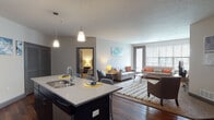 This single-floor model unit offers a thoughtfully designed open-concept layout that seamlessly connects the kitchen, dining, and living areas. The kitchen is outfitted with dark cabinetry, stainless steel appliances including a refrigerator, microwave, and gas range, and a central island with a light quartz countertop and double sink. Pendant lighting above the island adds a modern touch, while rich hardwood-style flooring anchors the space. The open living area is bright and welcoming, featuring large windows with horizontal blinds that allow ample natural light, and a glass sliding door leading to an outdoor balcony. The space accommodates generous seating arrangements and provides a comfortable, versatile setting. Multiple carpeted bedrooms offer a quiet retreat, each with windows for natural light and ample room for furnishings. The bathrooms are finished with dark vanity cabinetry, tile flooring, and well-lit mirrors; one includes a shower, and the other a full tub-shower combination. A dedicated in-unit laundry closet with a stacked washer and dryer provides added convenience. Throughout the unit, a neutral color palette and consistent dark-trimmed doors and accents create a cohesive, contemporary aesthetic.