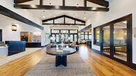 Located at 13600 Hilltop Groves Blvd in Clermont, FL, this single-story community clubhouse offers a generously sized, open-plan interior designed with a modern farmhouse aesthetic. Soaring vaulted ceilings with bold, dark-stained exposed beams define the architectural character of the space, creating a dramatic yet welcoming atmosphere throughout.

Warm-toned hardwood flooring flows continuously across the entire floor, unifying the various functional zones. The entry and reception area features a striking dark marble reception desk beneath a cluster of pendant lights, with the "Skybrone" brand name displayed prominently as a focal point. Large black-framed transom windows and glass panel doors line the walls, flooding the interior with natural light and offering outdoor views.

Multiple seating areas are thoughtfully arranged throughout the space. A central lounge features a generous sectional sofa accented with teal and yellow throw pillows, complemented by layered area rugs. A distinct fireside sitting area showcases a floor-to-ceiling white subway tile surround with a built-in fireplace, flanked by tall black-framed windows. Additional seating nooks include accent chairs and a built-in teal shelving unit. A dedicated workspace area with counter seating and a bar-height surface provides a functional co-working zone. The overall ambiance is vibrant, modern, and community-oriented.