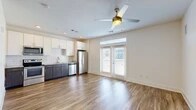 Located at 4454 Tropea Way in Jacksonville, Florida, this single-floor residence offers a clean, modern design with consistent wood-look flooring running throughout every room. The open-concept living and kitchen area serves as the heart of the home, anchored by a ceiling fan with light fixture and French doors that invite natural light and provide access to the outdoors. The kitchen is well-appointed with two-tone cabinetry — white upper cabinets paired with dark lower cabinets — granite-look countertops, a classic white subway tile backsplash, and a full suite of stainless steel appliances including a refrigerator, range, over-the-range microwave, and dishwasher. The bedroom is a comfortable, well-lit space featuring its own ceiling fan and a walk-in closet outfitted with wire shelving for organized storage. The bathroom is finished with wood-look tile flooring, a dark wood vanity with granite-look countertop, and a tub-shower combination with floor-to-ceiling white tile surround and a curved curtain rod. A hallway with louvered and paneled doors provides additional storage access. Bright white walls and recessed lighting contribute to the home's fresh, move-in-ready atmosphere throughout.