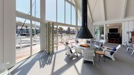 Located at 50 Barbaree Way in the sought-after waterfront community of Belvedere Tiburon, California, this single-level property offers a masterfully designed living experience with direct marina views and exceptional architectural detail throughout.

The space is defined by soaring vaulted ceilings with exposed white-painted beams and an abundance of natural light streaming through floor-to-ceiling glass walls. A dramatic covered lounge area features built-in warm wood banquette seating accented with navy and gray cushions, complemented by bistro tables and white Adirondack chairs arranged on large-format gray tile flooring. A striking suspended cone fireplace serves as a focal centerpiece, surrounded by casual seating that opens directly to the waterfront deck via folding glass panel doors, dissolving the boundary between interior and exterior.

The interior transitions seamlessly into a formal dining area with rich hardwood flooring, a statement chandelier, and industrial-style seating. A comfortable living room anchors the space with deep sectional sofas, a wall-mounted fireplace, curated artwork, and access to the water views beyond. Warm wood slatted railings and wide staircase detailing add refined architectural character throughout. The overall ambiance balances coastal relaxed living with sophisticated, contemporary design — ideal for both entertaining and everyday waterfront enjoyment.