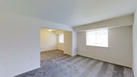 This single-floor residence offers a straightforward and functional layout designed for comfortable living. The main living area is spacious and open, finished with wall-to-wall carpeting in a neutral gray tone that runs seamlessly throughout the space. Smooth, white ceilings contribute to a clean and airy atmosphere, while crisp white baseboards frame the perimeter of each room. The walls are painted in a soft, warm neutral tone, providing a versatile backdrop well-suited to a variety of interior styles. Natural light enters generously through multiple windows fitted with horizontal blinds, brightening the interior and creating a welcoming ambiance. A defined archway or partial wall separates the main living area from an adjoining room, which features its own window and a doorway, offering a degree of separation while maintaining an open connection to the rest of the space. A wall-mounted heating vent is visible in the secondary room, indicating a central heating system. Flush-mount ceiling lighting provides additional illumination in the connected room. The overall layout is clean, uncluttered, and move-in ready, with a neutral palette that makes it easy to personalize.