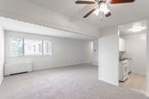 This single-floor unit offers a practical and comfortable layout across several well-defined spaces. The living area is generously sized, featuring soft wall-to-wall carpeting and a ceiling fan with integrated lighting. A decorative scalloped valance border divides the living space from a hallway, adding a subtle architectural detail. Natural light filters in through a window overlooking the exterior, enhancing the open feel of the room.

The kitchen is efficiently arranged with ample white cabinetry extending to the ceiling for generous storage. Dark speckled countertops provide a functional work surface alongside a white gas range and a stainless steel sink. The tile flooring in the kitchen offers easy maintenance and a clean contrast to the carpeted areas.

Two carpeted bedrooms are present, each bright and neutral in tone. Both rooms feature windows that allow natural light and views of the exterior, along with baseboard heaters. One bedroom includes a closet with bifold doors, providing dedicated storage.

The full bathroom is finished with warm beige wall tiles surrounding the tub-shower combination. A wood-toned vanity cabinet supports a white countertop with a single sink, complemented by a large mirror with built-in lighting above. Mosaic tile flooring completes the classic aesthetic of the bathroom.