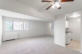 This single-floor unit offers a practical and comfortable layout across several well-defined spaces. The living area is generously sized, featuring soft wall-to-wall carpeting and a ceiling fan with integrated lighting. A decorative scalloped valance border divides the living space from a hallway, adding a subtle architectural detail. Natural light filters in through a window overlooking the exterior, enhancing the open feel of the room.

The kitchen is efficiently arranged with ample white cabinetry extending to the ceiling for generous storage. Dark speckled countertops provide a functional work surface alongside a white gas range and a stainless steel sink. The tile flooring in the kitchen offers easy maintenance and a clean contrast to the carpeted areas.

Two carpeted bedrooms are present, each bright and neutral in tone. Both rooms feature windows that allow natural light and views of the exterior, along with baseboard heaters. One bedroom includes a closet with bifold doors, providing dedicated storage.

The full bathroom is finished with warm beige wall tiles surrounding the tub-shower combination. A wood-toned vanity cabinet supports a white countertop with a single sink, complemented by a large mirror with built-in lighting above. Mosaic tile flooring completes the classic aesthetic of the bathroom.