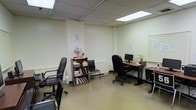 This single-floor office space offers a practical and organized environment designed to support day-to-day professional tasks. The interior features light-painted walls with a clean, neutral aesthetic, complemented by a drop ceiling with recessed fluorescent lighting panels that provide consistent, even illumination throughout the room. The flooring is finished in a smooth, light-toned surface that is easy to maintain and contributes to the bright overall feel of the space. Workstations are thoughtfully arranged to allow for individual focus, each equipped with ergonomic rolling chairs and desk surfaces accommodating computer monitors and essential work materials. A bookshelf unit serves as a central organizational element, neatly storing books, binders, and decorative touches such as a small potted plant and floral arrangement, adding a modest warmth to the utilitarian setting. Window blinds along one wall allow for natural light control, offering flexibility in managing the room's brightness. The space also features a compact secondary seating area with a side chair, providing additional flexibility for collaborative or consultative use. Overall, this office strikes a balance between efficiency and comfort, making it a well-suited environment for focused professional or administrative work.