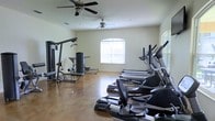 This fitness center offers a comprehensive workout environment designed to accommodate a range of exercise needs. The space is outfitted with multiple cardio machines, including treadmills and ellipticals, as well as a multi-station strength training unit, providing users with the tools for a full-body workout. The room features a polished concrete floor, which is both durable and easy to maintain, and is finished with neutral walls that create a clean, focused atmosphere. Natural light filters in through an arched window fitted with horizontal blinds, brightening the interior and contributing to an open, airy feel. Two ceiling fans are mounted overhead, ensuring consistent air circulation throughout the workout space and keeping the environment comfortable during use. The layout is thoughtfully arranged, with cardio equipment positioned along one side and strength machines along the other, allowing for efficient use of the available floor space. The overall design is functional and practical, emphasizing accessibility and ease of movement throughout the room.