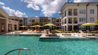 This amenity space is anchored by a stunning resort-style pool featuring crystal-clear turquoise water, an in-pool sun shelf with lounge chairs, and a surrounding deck appointed with lime-green market umbrellas and additional seating areas. The pool area is framed by lush landscaping and a decorative fence, with a multi-story building providing an attractive backdrop beneath open, blue skies.

The fitness center is thoughtfully divided into distinct workout zones. A strength-focused area houses a multi-station cable machine, power rack with bench, rowing ergometer, and a full dumbbell rack with weights neatly arranged for easy access. Medicine balls, battle ropes, resistance bands, and stability balls round out the functional training equipment. Floor-to-ceiling mirrors visually expand the space, while ceiling fans promote airflow throughout. Herringbone-patterned, multi-tone blue carpet tiles cover the floors.

A dedicated cardio room adjoins the main fitness floor, featuring treadmills positioned before large windows that invite natural light and outdoor views. A wall-mounted television provides entertainment during workouts. Together, these amenities create a comprehensive, well-designed environment suited for both relaxation and active lifestyles.