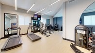 Located at 6180 Rock Island Road in Tamarac, Florida, this fitness center offers a comprehensive workout facility within a well-designed, single-floor space. The room features a clean, modern aesthetic with light wood-style flooring, crisp white ceilings, and bold blue-grey accent walls that create an energizing atmosphere.

The cardio zone is equipped with treadmills, elliptical machines, and stationary bikes, all positioned beneath sleek linear LED lighting that runs across the ceiling. Wall-mounted flat-screen TVs are strategically placed throughout the space, providing entertainment during workouts.

The strength training area is equally well-appointed, featuring a multi-station cable machine, a variety of selectorized weight machines, and a dedicated free weights section with a full rack of dumbbells and kettlebells arranged along the wall. Adjustable benches and additional resistance machines round out the equipment offerings.

Large mirrors are incorporated into the layout, enhancing the sense of openness and allowing users to monitor their form. Natural light filters in through several windows, while recessed and linear lighting ensures the space remains bright throughout. A glass partition offers a visual connection to an adjacent lounge or common area, adding a sense of community to the fitness environment.