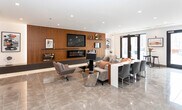 Located at 19 E 19th St in Bayonne, NJ, this single-floor amenity space offers a refined, contemporary design with high-end finishes throughout. The main lounge area makes an immediate impression with its polished large-format tile flooring, recessed lighting, and a dramatic wood-paneled feature wall housing a wall-mounted flat-screen TV and a sleek linear electric fireplace with ambient underlighting. Comfortable seating arrangements — including plush sofas and modern accent chairs — create an inviting atmosphere suited for both relaxation and social gatherings. Curated artwork adorns the walls, adding a cultivated aesthetic to the space. Adjacent to the lounge, a well-appointed dining and bar area provides additional seating at extended countertops and bar-height stools with warm pendant lighting overhead. The fully equipped kitchenette features dark cabinetry, marble-look quartz countertops and backsplash, a stainless steel sink, undercounter appliances, and open display shelving for décor. The open layout allows the lounge, dining, and kitchen areas to flow seamlessly together, making the space highly functional and visually cohesive. The overall ambiance is polished and modern, ideal for community gatherings or events.