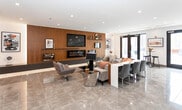 Located at 19 E 19th St in Bayonne, NJ, this single-floor amenity space offers a refined, contemporary design with high-end finishes throughout. The main lounge area makes an immediate impression with its polished large-format tile flooring, recessed lighting, and a dramatic wood-paneled feature wall housing a wall-mounted flat-screen TV and a sleek linear electric fireplace with ambient underlighting. Comfortable seating arrangements — including plush sofas and modern accent chairs — create an inviting atmosphere suited for both relaxation and social gatherings. Curated artwork adorns the walls, adding a cultivated aesthetic to the space. Adjacent to the lounge, a well-appointed dining and bar area provides additional seating at extended countertops and bar-height stools with warm pendant lighting overhead. The fully equipped kitchenette features dark cabinetry, marble-look quartz countertops and backsplash, a stainless steel sink, undercounter appliances, and open display shelving for décor. The open layout allows the lounge, dining, and kitchen areas to flow seamlessly together, making the space highly functional and visually cohesive. The overall ambiance is polished and modern, ideal for community gatherings or events.