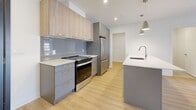 Located at 17 Place de la Triade in Pointe-Claire, Quebec, this modern 2-bedroom, 2-bathroom unit offers a well-designed open-concept layout with clean, contemporary finishes throughout. Light hardwood flooring runs through the main living areas, complementing the bright white walls and recessed lighting that create a fresh, airy atmosphere.

The kitchen serves as the centerpiece of the main living space, featuring a large island with a white countertop, integrated sink, stainless steel appliances including a refrigerator and dishwasher, and a combination of warm wood-toned upper cabinetry and grey lower cabinets with a tile backsplash. Pendant lights hang above the island, adding a stylish touch. The kitchen flows seamlessly into the combined dining and living area, which is bathed in natural light from a sliding glass door that opens onto a private balcony with open exterior views.

Both bedrooms are well-proportioned, featuring hardwood flooring and ample closet space. The primary bedroom includes sliding door access to the balcony. The two bathrooms are finished with large-format beige floor tiles and warm wood-toned vanity cabinetry with white countertops. A dedicated in-unit laundry room houses a full-size front-loading washer and dryer. Three closets and a welcoming entrance complete this thoughtfully designed residence.