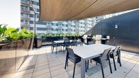 Located at 1140 Rue Wellington in Montréal's dynamic Griffin/Griffintown neighbourhood, this rooftop terrace is a thoughtfully designed outdoor amenity space offering a compelling mix of relaxation and al fresco dining. The terrace is finished with large-format stone-effect tiles that lend a clean, contemporary aesthetic throughout. Lush greenery lines the perimeter in sleek, dark-toned planters, creating a sense of privacy while softening the urban surroundings with vibrant tropical foliage. The space is divided into two distinct zones: a social lounge area featuring a generous sectional sofa and a round coffee table, ideal for casual gatherings, and a dedicated dining area outfitted with multiple rectangular tables and modern chairs to accommodate larger groups. Overhead sail shade canopies provide comfortable shelter from the sun across the dining section. A BBQ station with multiple grills is conveniently positioned nearby, making this terrace well-equipped for outdoor cooking and entertaining. Panoramic views of the Montréal skyline, including its iconic downtown towers, can be enjoyed from the terrace, offering a striking urban backdrop. The open-air design maximizes natural light and airflow, creating a bright and inviting atmosphere throughout the day.