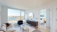 Located at 20 Hallets Point in Astoria, Queens, this one-bedroom, one-bathroom residence offers a thoughtfully designed modern interior with sweeping waterfront views of the East River and the iconic Hell Gate Bridge. The open-concept living area is anchored by expansive floor-to-ceiling windows that flood the space with natural light while framing breathtaking views of the river and surrounding landscape. Light-toned hardwood floors run throughout, complementing the clean, contemporary aesthetic. The kitchen features a large waterfall-edge island with bar seating, a combination of white upper cabinetry and warm wood lower cabinetry, stone backsplash, stainless steel appliances, and a gas range — ideal for both everyday cooking and entertaining. The living room connects seamlessly to the kitchen and offers generous space for lounging and dining. The bedroom is a serene retreat highlighted by a large corner window with panoramic river views and ample closet storage with dual built-in closets. The bathroom is finished with large-format white tiles, a deep soaking tub, a floating wood vanity, and a backlit mirror, creating a spa-like atmosphere. A light-filled entryway hallway with recessed lighting completes this well-appointed home.