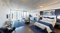 This thoughtfully designed single-floor studio unit showcases a seamless open-concept layout that maximizes both functionality and visual appeal. The space is anchored by dramatic floor-to-ceiling windows that flood the interior with natural light while framing sweeping panoramic city views, creating a striking connection between indoors and the urban landscape beyond.

The exposed concrete ceiling lends an industrial-chic character throughout, complemented by warm hardwood flooring that unifies the living, sleeping, and kitchen areas. The sleeping area features a bold navy accent wall, providing a defined focal point within the open plan, alongside a closet for storage.

The kitchen is well-appointed with stainless steel appliances, including a side-by-side refrigerator, dishwasher, electric range, and microwave. White shaker cabinetry extends to the ceiling, offering generous storage, while granite countertops add a refined finish. An in-unit stacked washer and dryer provide added convenience.

The bathroom is finished with light tile flooring, a granite-topped vanity with white cabinetry, and a shower-tub combination. The overall design palette of navy, white, and gold accents conveys a cohesive, upscale aesthetic throughout the space.