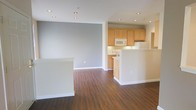 This single-floor space showcases a thoughtfully designed open-concept layout that connects the living area and kitchen in a cohesive, flowing arrangement. Rich hardwood-style flooring extends throughout, adding warmth and continuity to the space. The living area is spacious and versatile, framed by neutral gray walls and crisp white baseboards that create a clean, modern aesthetic.

The kitchen is positioned toward the rear and features light wood cabinetry that extends to the upper walls, providing ample storage. White appliances, including a refrigerator, microwave, and range, are neatly integrated into the layout. A kitchen peninsula serves as a functional divider between the cooking and living spaces, offering additional counter space and a natural transition point between the two areas.

Recessed lighting is installed throughout the ceiling, ensuring even, consistent illumination across the entire space. The open floor plan and neutral color palette create a bright and airy ambiance that lends itself well to a variety of furniture arrangements and interior styles. The overall design emphasizes practicality and simplicity, making efficient use of the available space.