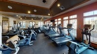 This fitness center is thoughtfully designed to support a comprehensive workout experience within a single open floor layout. The space features a distinctive architectural character, with exposed wooden beam accents framing a vaulted ceiling finished in a deep charcoal tone, complemented by warm terracotta-colored accent walls and recessed lighting throughout.

Natural light floods the interior through multiple large, wood-trimmed windows lining the perimeter walls, offering views of the outdoors and creating a bright, energizing atmosphere. Durable rubber flooring covers the entire workout area, providing both safety and comfort underfoot.

The cardio section is generously appointed with a row of treadmills, elliptical trainers, stationary bikes, and stair-climbing machines, many equipped with individual monitors for entertainment or workout tracking. Full-length mirrors along one wall visually expand the space and assist with form during exercise.

The strength training area includes a multi-function cable and weight machine, a leg press, a functional trainer, and additional resistance equipment, offering a versatile range of training options. TRX suspension straps are also mounted and available for bodyweight training.

French-style glass doors provide access to what appears to be an outdoor area, further enhancing ventilation and connectivity to the surrounding environment. Overall, the space balances functional fitness amenities with warm, inviting design elements.