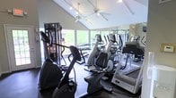 This fitness center offers a comprehensive workout environment within a single-level layout topped by striking vaulted ceilings that lend an open, airy feel to the space. Large windows line the walls, flooding the room with natural light and providing views of surrounding greenery, creating an inviting atmosphere for exercise. A ceiling fan overhead aids in air circulation throughout workouts.

The facility is outfitted with a strong selection of Matrix-brand cardio machines, including treadmills, elliptical trainers, and stationary bikes, arranged to maximize usable floor space while maintaining comfortable spacing between equipment. A dedicated free weights area features a full dumbbell rack along one wall, catering to strength training needs. A large wall mirror enhances the sense of depth and allows users to monitor their form during workouts.

The flooring is dark rubberized material, providing both durability and safety underfoot. Track lighting complements the natural light, ensuring the space remains well-illuminated at all hours. The overall ambiance is clean, modern, and functional, designed to support a wide range of fitness activities in a comfortable, well-maintained environment.