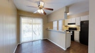 This single-story space showcases an open-concept layout that seamlessly connects the living area and kitchen, creating a cohesive and functional floor plan. The living area features rich, dark hardwood-style flooring that contrasts attractively with the warm beige walls and bright white trim. A ceiling fan with an integrated light fixture is centrally positioned, providing both air circulation and ambient lighting. Large sliding glass doors dressed with vertical blinds span nearly the full width of the living area, flooding the space with abundant natural light and offering views of the surrounding outdoor greenery.

The kitchen is defined by a raised breakfast bar counter that serves as a natural divider between the two spaces while maintaining the open feel. White cabinetry lines the upper portion of the kitchen walls, complemented by a black range and an overhead ventilation hood. A window above the kitchen counter allows additional natural light into the cooking area. The textured ceiling adds a traditional finishing detail throughout. Overall, the space presents a practical and inviting atmosphere well-suited for comfortable everyday living.