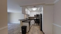This single-floor space offers a clean, modern aesthetic highlighted by thoughtful design choices and quality finishes throughout. The kitchen serves as a standout feature, designed with functionality and style in mind. It is outfitted with a full suite of stainless steel appliances, including a gas range with oven, over-the-range microwave, and a black refrigerator, providing a sleek contrast against the bright white shaker-style cabinetry. Dark countertops extend across the workspace and wrap around a peninsula that opens toward the adjacent living area, creating a natural flow between cooking and living spaces. A double-basin sink with a modern faucet is neatly positioned along the peninsula, adding to the kitchen's practicality. The kitchen benefits from a recessed tray ceiling with flush-mount lighting, lending an elevated feel to the space. Crown molding adds a refined architectural detail that carries through to the adjoining room. Wood-style plank flooring runs throughout, offering warmth and durability underfoot, while neutral wall tones keep the space feeling bright and open. The open-concept layout between the kitchen and living area enhances the overall sense of spaciousness, making this a well-designed and comfortable living environment.