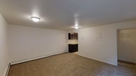 This single-floor unit offers a straightforward, functional layout designed for efficient use of space. The main living area features wall-to-wall carpeting in a neutral tan tone, providing a comfortable underfoot feel. White walls and a textured ceiling create a clean, simple backdrop, while two flush-mount ceiling light fixtures ensure consistent overhead illumination throughout the space.

A baseboard heater runs along the far wall of the living area, indicating an electric heating system. A wall panel, likely housing electrical controls or a utility access point, is visible on the adjacent wall.

Directly accessible from the living area, the kitchen is distinguished by dark espresso-toned lower cabinetry that contrasts with the lighter surroundings. The kitchen appears compact yet functional, with cabinetry providing ample storage. The transition between the carpeted living space and the kitchen is clearly defined by the change in flooring.

Overall, the space maintains a neutral, understated aesthetic with minimal ornamentation, making it well-suited for a variety of furnishing styles and practical everyday use.