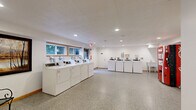 Located at 600 North Lilac Drive in Golden Valley, Minnesota, this shared laundry facility offers a clean, well-organized space designed for convenience and functionality. The room features durable speckled aggregate flooring throughout, recessed lighting in a textured ceiling, and several windows along one wall that allow natural light to filter in while providing views of the surrounding greenery outside.

The facility is equipped with multiple front-loading washing machines and dryers arranged in rows, offering ample capacity for residents. Each machine features card or coin-operated controls, making transactions straightforward. A utility sink is available along one wall, providing added convenience for handwashing or pre-treating garments.

An additional area within the space includes garment racks and a rolling laundry cart, offering practical support for sorting and transporting clothes. A Coca-Cola vending machine is positioned in the room, adding a convenient amenity for users during their visit.

Seating is available in the form of decorative wrought-iron chairs, accompanied by framed wall art, lending a touch of comfort and character to the otherwise utilitarian space. A glass-paned exterior door provides direct outdoor access and adds to the brightness of the room. Safety features including clearly marked exit signage and a fire alarm system are also present throughout the facility.