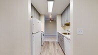 Located at 9300 W Mall Dr in Everett, Washington, this single-floor unit offers four bedrooms and two bathrooms with a thoughtfully laid-out floor plan. The galley-style kitchen is a standout feature, lined on both sides with soft gray shaker cabinetry extending floor to ceiling, providing generous storage. Marble-patterned countertops complement the cabinetry, and the kitchen is equipped with a full-size refrigerator, electric range, and dishwasher. A skylight-style overhead fixture and ceiling fan at the adjoining hallway brighten the space, while light wood-look flooring runs throughout the kitchen. The living room is carpeted and generously proportioned, featuring a sliding glass door that opens to an outdoor area, allowing natural light to filter in. Each of the four bedrooms is finished with plush carpeting and neutral wall tones; the bedrooms feature windows with views of lush greenery, contributing to a calm, natural ambiance. The primary bedroom offers a comfortable footprint with closet access. Both bathrooms are finished with wood-look tile flooring and gray vanity cabinetry with stone-topped counters; one bathroom includes a full soaking tub and shower combination. A central hallway connects the living areas to the bedrooms, providing a practical and organized flow throughout the home.