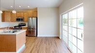 This single-floor residence offers a thoughtfully designed layout that balances comfort and functionality. The kitchen serves as a standout feature, equipped with warm honey-toned wood cabinetry, stainless steel appliances including a gas range, built-in microwave, and French door refrigerator, and a central island with a built-in sink and pendant lighting overhead. Light wood-style flooring extends through the kitchen and adjoining dining area, which benefits from a large sliding glass door that invites abundant natural light and outdoor views. The open layout connects the kitchen seamlessly to the living areas, creating an airy, cohesive flow throughout the space. The bedrooms are finished with plush neutral carpeting and feature generous walk-in closets with built-in shelving and hanging rods, offering ample storage. Windows in the bedrooms frame views of greenery outside, enhancing the sense of tranquility. The full bathroom is finished with light tile flooring, a soaking tub, and a window providing natural ventilation, while a convenient half bathroom features wood-style flooring and clean, minimal fixtures. A dedicated laundry area is also present within the unit. Throughout, neutral wall tones and white trim create a clean, welcoming ambiance.