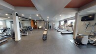 This fitness center at 2710 N Proctor St in Tacoma, Washington, offers a thoughtfully organized, single-level space designed to accommodate a broad range of workout needs. The interior features durable rubber flooring throughout, providing both safety and comfort during exercise. Recessed lighting illuminates the space evenly, complemented by natural light streaming in through multiple windows along one wall, which also offer views of the surrounding greenery.

The cardio zone is equipped with multiple treadmills, each paired with wall-mounted flat-screen televisions for entertainment during workouts. The strength training area includes a multi-station cable machine, a rack of dumbbells arranged along a floor-to-ceiling mirror, and a weight bench, giving users a comprehensive free-weight and resistance training setup. A dedicated functional fitness area contains stability balls in various sizes, resistance bands, foam rollers, and yoga mats stored on an organized rack, making it ideal for stretching, core work, and flexibility training.

Accent details include warm wood-panel wall features and terracotta-toned ceiling insets that add visual character to the otherwise neutral gray palette. A water dispenser and storage shelving are conveniently positioned within the space. A seating area with a cushioned chair provides a spot to rest between sets, rounding out this well-appointed fitness facility.