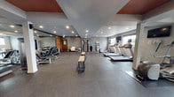This fitness center at 2710 N Proctor St in Tacoma, Washington, offers a thoughtfully organized, single-level space designed to accommodate a broad range of workout needs. The interior features durable rubber flooring throughout, providing both safety and comfort during exercise. Recessed lighting illuminates the space evenly, complemented by natural light streaming in through multiple windows along one wall, which also offer views of the surrounding greenery.

The cardio zone is equipped with multiple treadmills, each paired with wall-mounted flat-screen televisions for entertainment during workouts. The strength training area includes a multi-station cable machine, a rack of dumbbells arranged along a floor-to-ceiling mirror, and a weight bench, giving users a comprehensive free-weight and resistance training setup. A dedicated functional fitness area contains stability balls in various sizes, resistance bands, foam rollers, and yoga mats stored on an organized rack, making it ideal for stretching, core work, and flexibility training.

Accent details include warm wood-panel wall features and terracotta-toned ceiling insets that add visual character to the otherwise neutral gray palette. A water dispenser and storage shelving are conveniently positioned within the space. A seating area with a cushioned chair provides a spot to rest between sets, rounding out this well-appointed fitness facility.