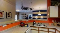 This single-floor space makes a strong visual impression through its bold and energetic interior design choices. The kitchen serves as a central hub, featuring a double-basin stainless steel sink, ample counter space along an L-shaped layout, and white cabinetry beneath warm-toned countertops. A standout wrought iron pot rack hangs from the ceiling, providing both functional storage and an industrial design accent that adds character to the space.

The kitchen opens directly into the living area, creating a seamless, open-concept flow. A striking navy blue and cream horizontal-striped accent wall anchors the living space, offering a bold nautical aesthetic that contrasts with the vibrant orange lower wall panels visible in the kitchen. Pendant lighting — including a classic cone-shaped pendant over the dining or transitional area — adds warmth and elegance.

The living area features neutral walls complemented by a decorative wall-mounted art arrangement, a tall navy blue vase as a decorative accent, and softly lit table lamps that contribute to a cozy ambiance. Natural light filters in through a window dressed with curtains, further brightening the space. Together, the design elements create a lively yet inviting atmosphere that balances bold color choices with functional, well-organized living areas.