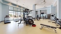 Located at 211 River Landing Dr in Charleston, South Carolina, this generously sized fitness center offers a thoroughly outfitted exercise environment across a single open floor. The space impresses immediately with soaring double-height ceilings, soft blue-gray walls, and a striking decorative mosaic tile accent wall that adds a modern, polished aesthetic. Large floor-to-ceiling windows along one side of the room flood the interior with natural light and provide outdoor views of the surrounding landscape, creating an open and energizing atmosphere. Ceiling fans and recessed lighting ensure consistent comfort and visibility throughout. The exercise floor features durable, light-toned flooring and is comprehensively equipped with a wide selection of free weights, including multiple dumbbell racks and barbells, adjustable benches, cable and multi-station cable machines, a functional trainer, treadmills, ellipticals, upright and recumbent bikes, rowing machines, and medicine balls. Full-length mirrors line one wall, enhancing the sense of space while supporting proper form during training. A dedicated entry area includes built-in wooden cubbies for personal belongings, a water station, and a wall-mounted flat-screen television, complementing the additional TV mounted in the main workout zone.