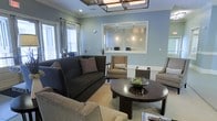 This single-floor space presents a thoughtfully designed interior centered around a comfortable and visually cohesive lounge area. The walls are painted in a soft, calming blue-gray tone, complemented by crisp white wainscoting that adds a classic architectural detail throughout. A statement geometric chandelier anchors the ceiling, providing both ambient and focused lighting above the seating area. Crown molding further refines the aesthetic, lending a polished finish to the room.

The seating arrangement consists of a dark charcoal sofa accented with decorative pillows, paired with neutral-toned accent chairs that create an inviting, balanced layout. A dark-stained round coffee table serves as a focal point, topped with a decorative centerpiece. A textured area rug grounds the grouping and adds warmth underfoot.

A prominent interior window with a mosaic-tiled surround opens to what appears to be a reception or service counter beyond, suggesting a professional or hospitality-oriented function for this space. Natural light filters in through curtained windows, enhancing the room's welcoming atmosphere. The overall ambiance is refined, comfortable, and well-suited for receiving guests.