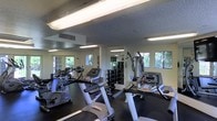 This fitness center is a fully equipped exercise facility designed to support a wide range of workout routines. The space is outfitted with multiple cardio machines, including treadmills and stationary bikes, arranged to allow comfortable movement throughout the room. A dedicated free weights section features a full dumbbell rack, providing options for strength training at various levels. A multi-station cable and resistance machine adds further versatility for full-body workouts. The room is lined with mirrors along one wall, enhancing the sense of space and allowing users to monitor their form during exercise. Overhead fluorescent lighting ensures the interior is consistently well-lit, while several windows along the perimeter walls invite natural light and offer views of surrounding greenery, creating a pleasant and motivating atmosphere. The flooring is dark-toned and gym-appropriate, providing durability and support underfoot. The textured ceiling and neutral wall tones give the space a clean, functional aesthetic. The HVAC vent integrated into the ceiling supports climate control, ensuring a comfortable environment year-round. Overall, this fitness center is a practical, well-appointed space suited for a comprehensive exercise regimen.