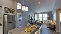 This well-appointed single-floor residence showcases a thoughtfully designed open-concept layout that seamlessly connects the kitchen, dining, and living areas into one cohesive and inviting space. Vaulted ceilings soar overhead, amplifying the sense of openness and volume throughout the main living area. A trio of decorative pendant lights hangs above the dining area, adding a warm, contemporary focal point to the space.

The kitchen features sleek countertops and stainless steel appliances, flowing naturally into the dining space, which is set with a stylish table and modern seating. The living area is anchored by a media console with an entertainment center, flanked by large windows that flood the room with abundant natural light and offer views of the surrounding exterior.

Neutral gray tones dominate the walls and furnishings, accented by pops of yellow in the décor and dishware, creating a cheerful and modern aesthetic. Hardwood-style flooring runs throughout the main living spaces, adding warmth and continuity. A bedroom visible through an open doorway continues the calm, neutral palette with soft, coordinating furnishings. The overall ambiance is bright, clean, and contemporary, offering a comfortable and functional living environment.
