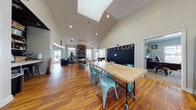 The Edge @ Kutz is a well-appointed single-floor space designed to serve a variety of functions, blending communal gathering areas with dedicated workspaces. The main hall is the centerpiece of the layout, distinguished by dramatic vaulted ceilings with a skylight that floods the room with natural light. Warm-toned hardwood flooring extends throughout, complementing the bright, neutral walls and creating an inviting atmosphere. A striking floor-to-ceiling stone fireplace anchors one end of the main hall, flanked by large windows that allow ample natural light to pour in. The open layout accommodates multiple seating configurations, including long communal tables with teal metal chairs and smaller groupings with white metal chairs, making it suitable for collaborative work, events, or casual gatherings. A snack or refreshment counter lines one wall, adding to the space's multipurpose appeal. Recreational amenities are also present, including a ping-pong table and what appears to be an air hockey table. A dedicated private office is accessible from the main area, featuring carpeted flooring, crown molding, multiple windows with blinds, and ample storage shelving. The overall design strikes a balance between functional utility and a welcoming, energetic ambiance suited for community engagement and productivity.